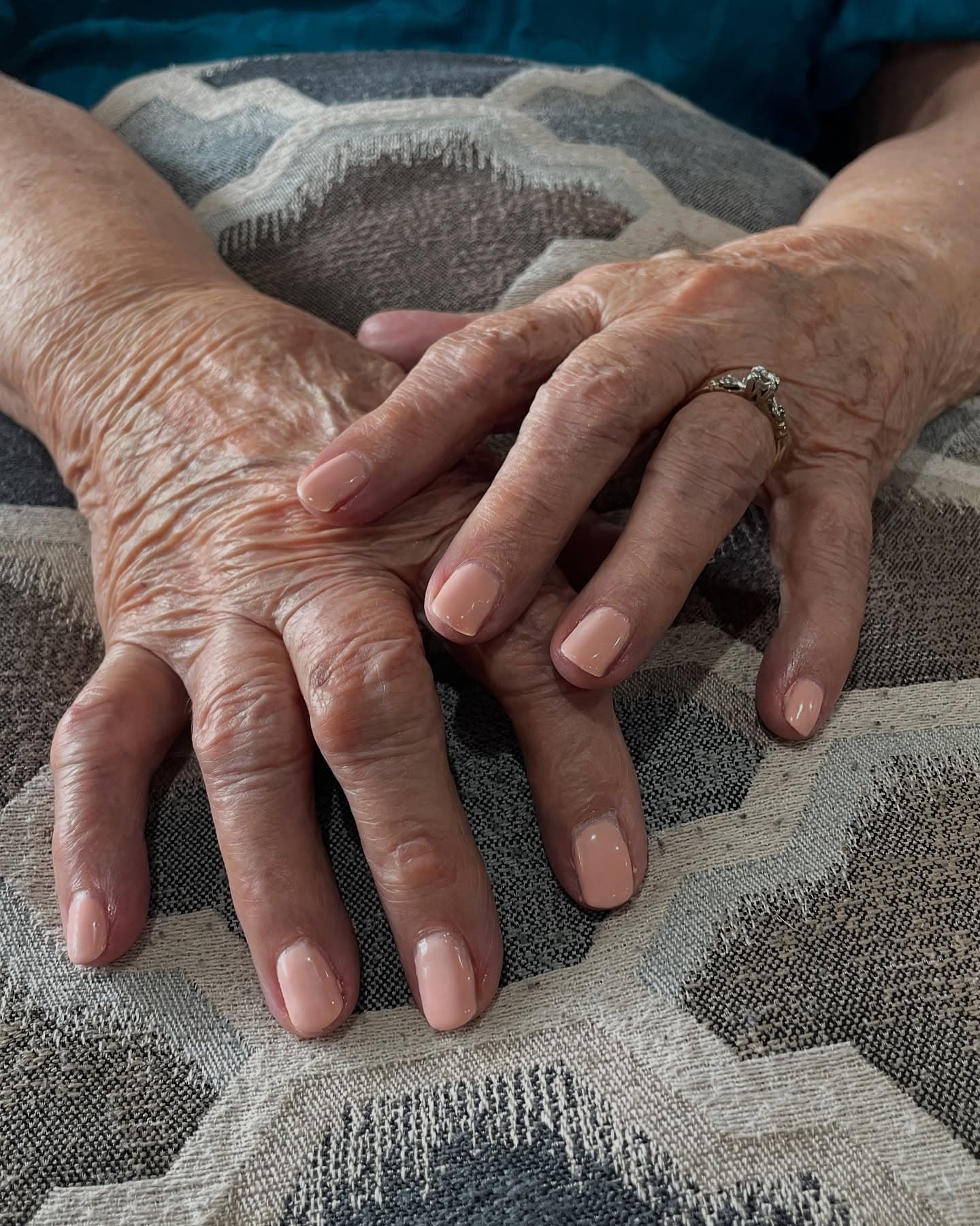 An elderly client's short rounded nails painted in a soft peachy nude, hands resting gently on a patterned cushion