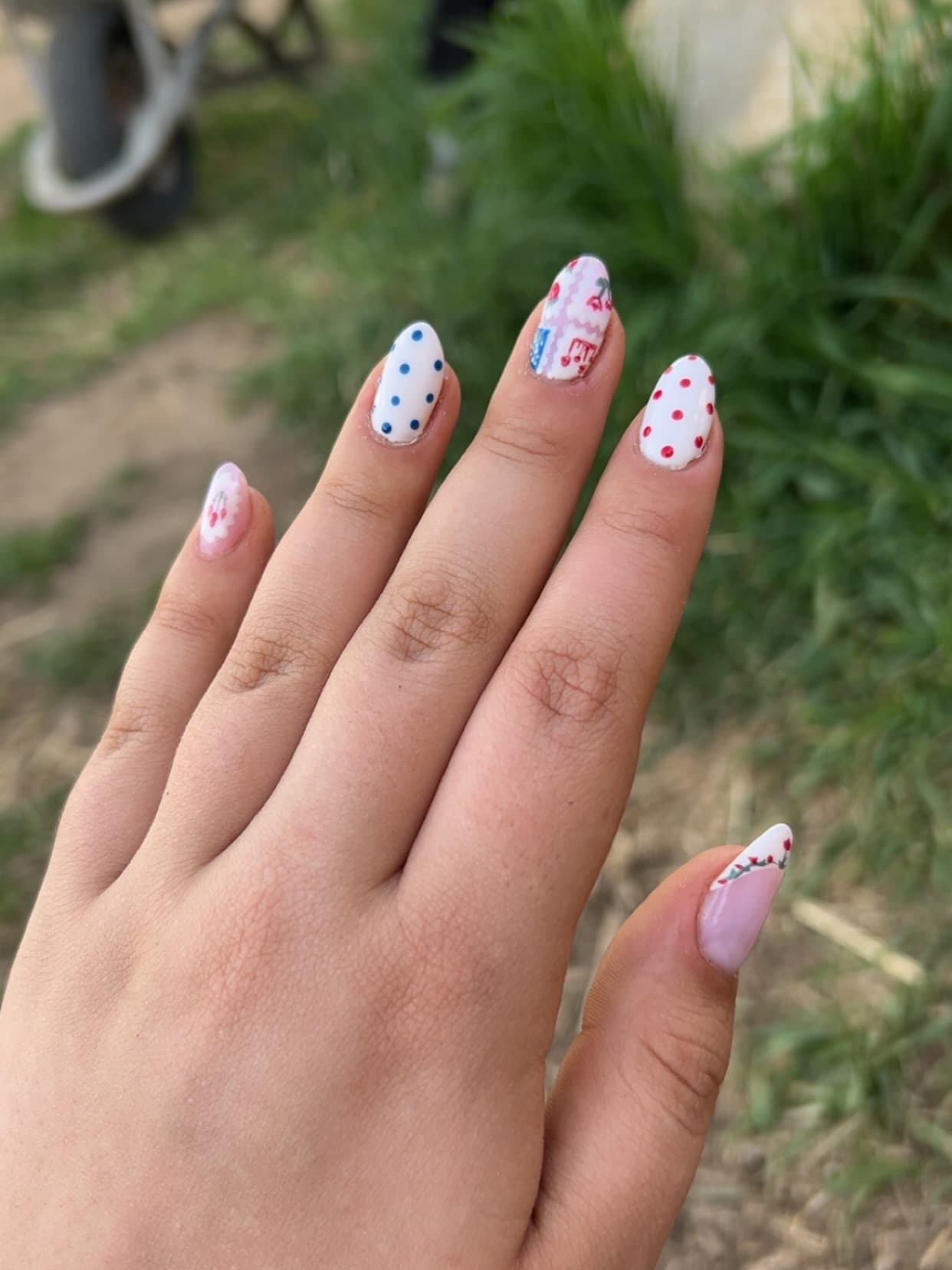 Almond nails held up outdoors, mixing polka dots on white with tiny cherries, pink ric-rac trim and a rosebud tip on the thumb