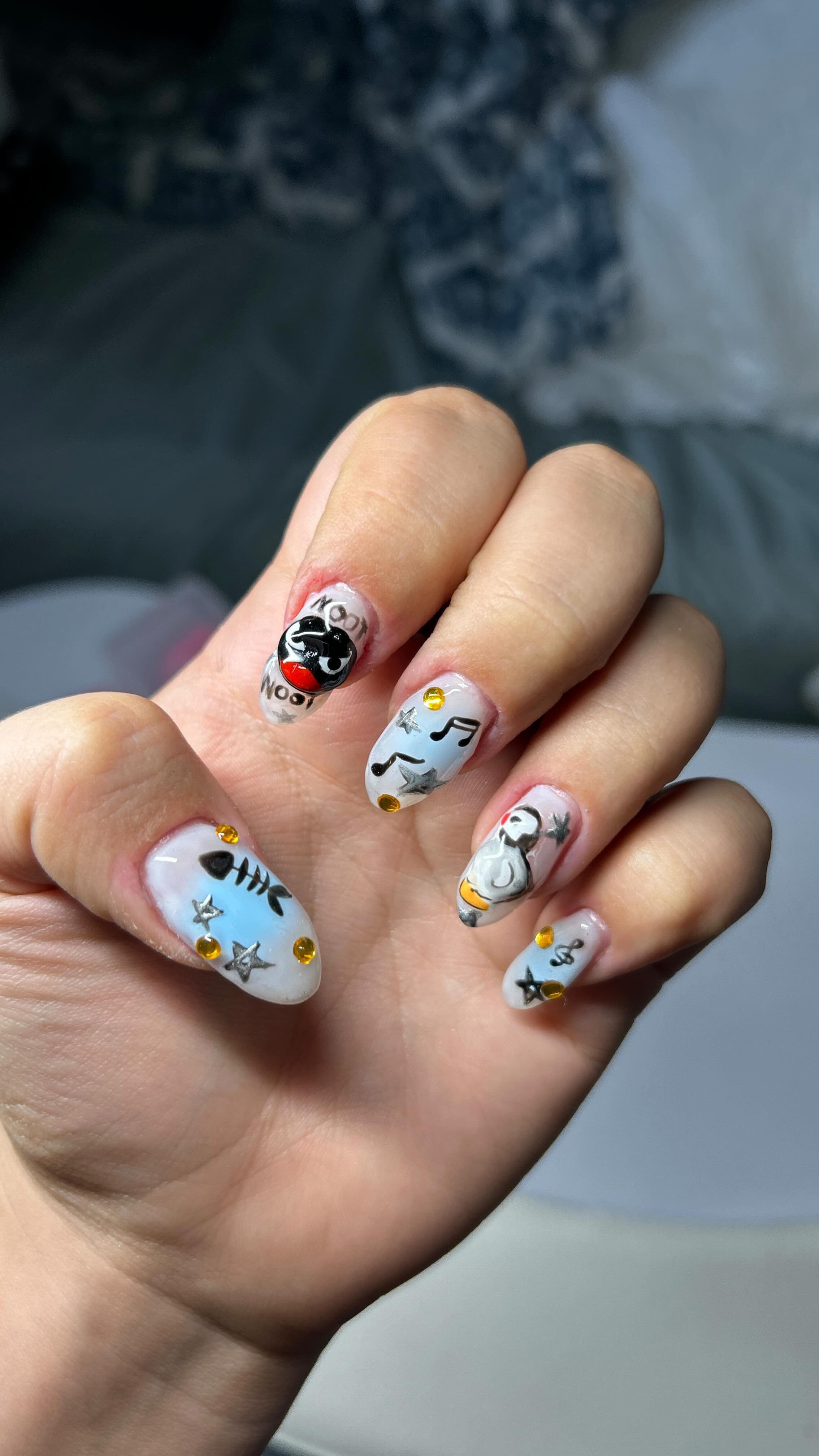 Long almond nails in milky blue with a hand-painted Pingu penguin, noot-noot lettering, fish bones, music notes, silver stars and amber gems