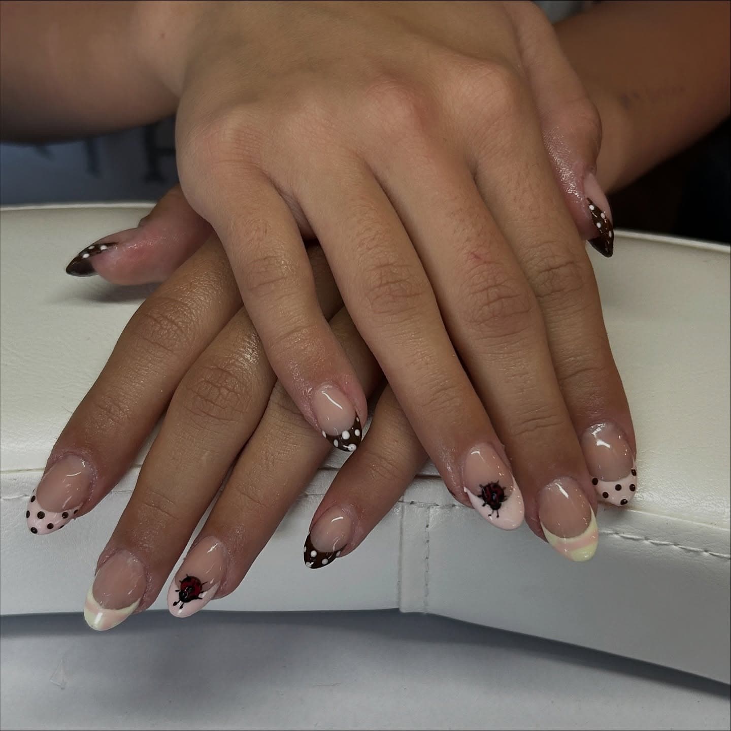 Almond nails in sheer nude with mismatched French tips of chocolate polka dots, butter yellow and blush pink, finished with tiny ladybug accents