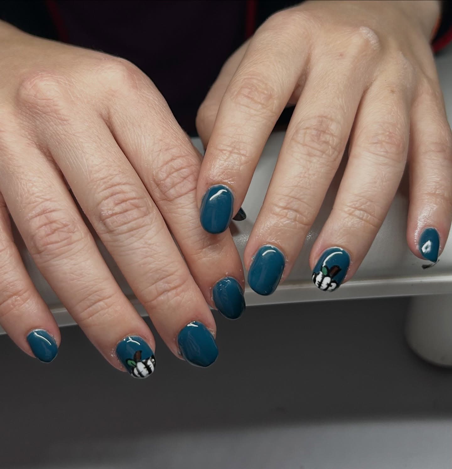 Short round nails in deep teal with a hand-painted white pumpkin accent on each hand