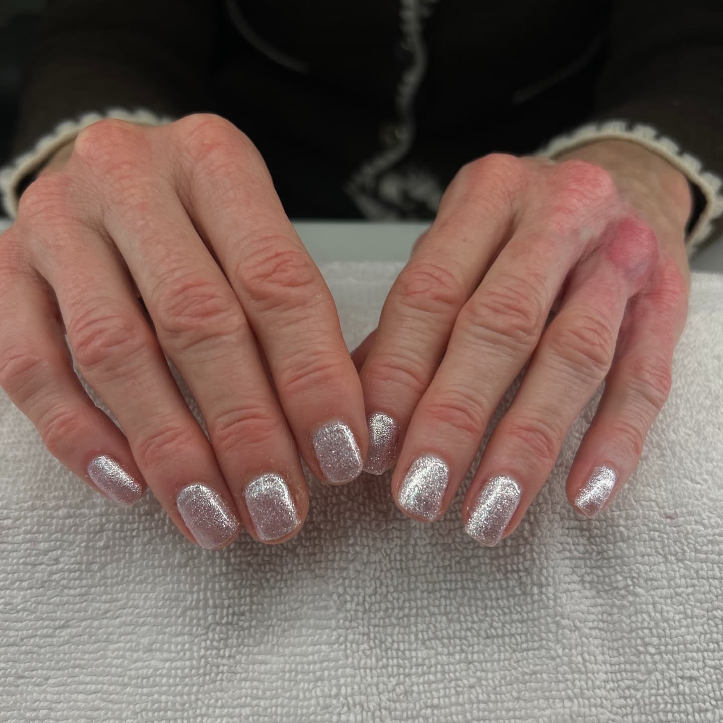 An elderly client's short square nails in sparkling champagne-silver glitter resting on a white towel