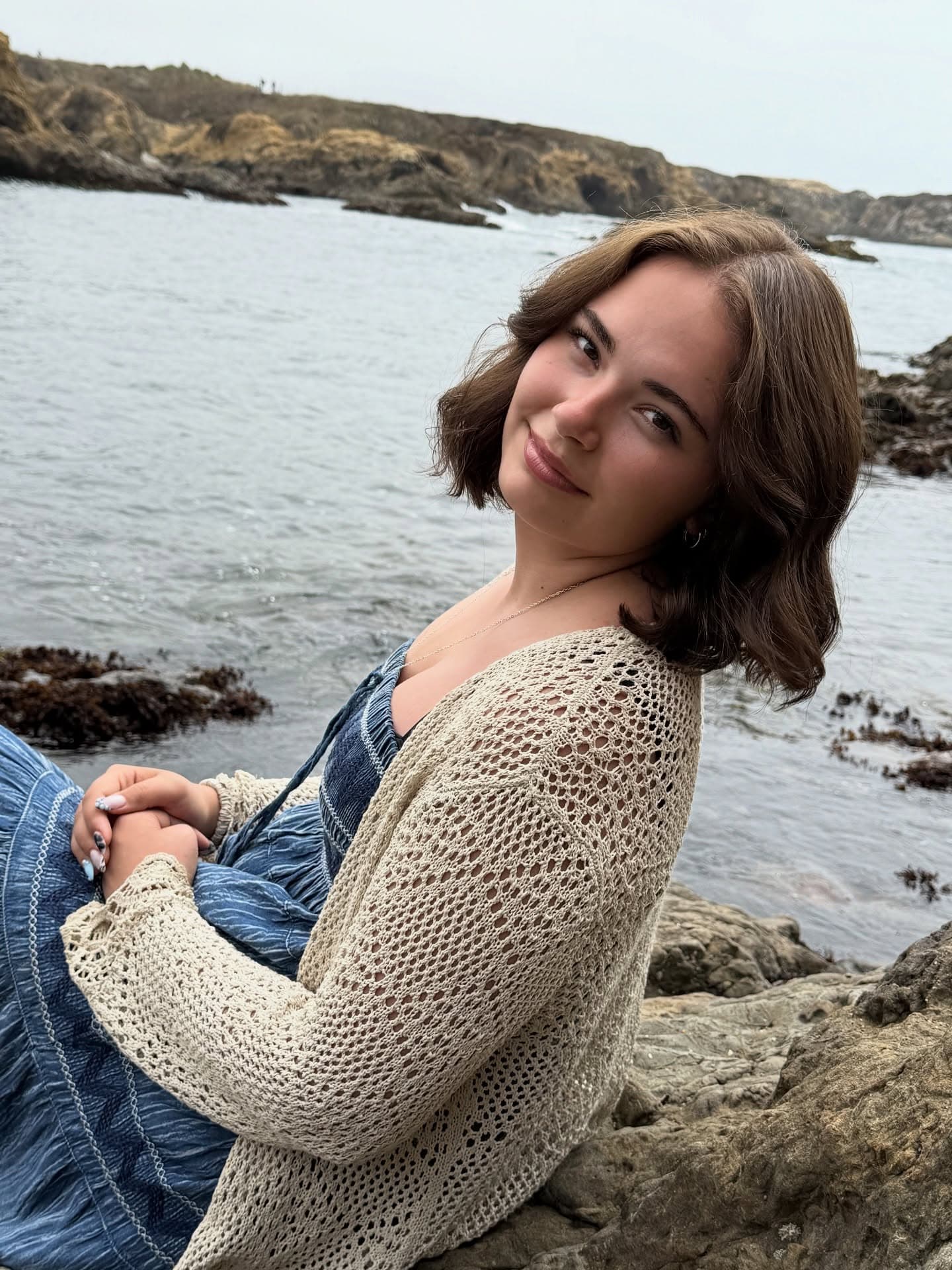 A client with a wavy bob sits on seaside rocks, her folded hands showing a powder-blue and patterned manicure